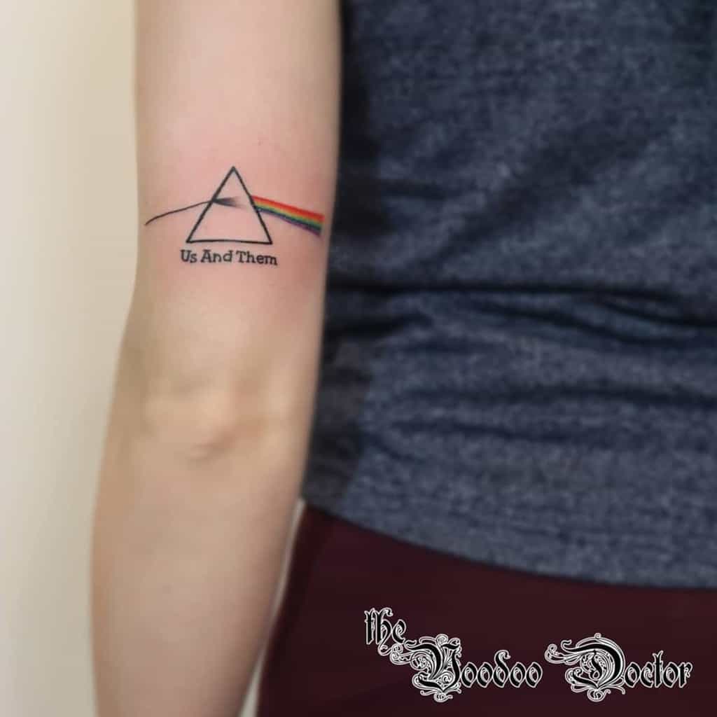 Tattoo of a prism with rainbow and text "Us And Them" on an arm