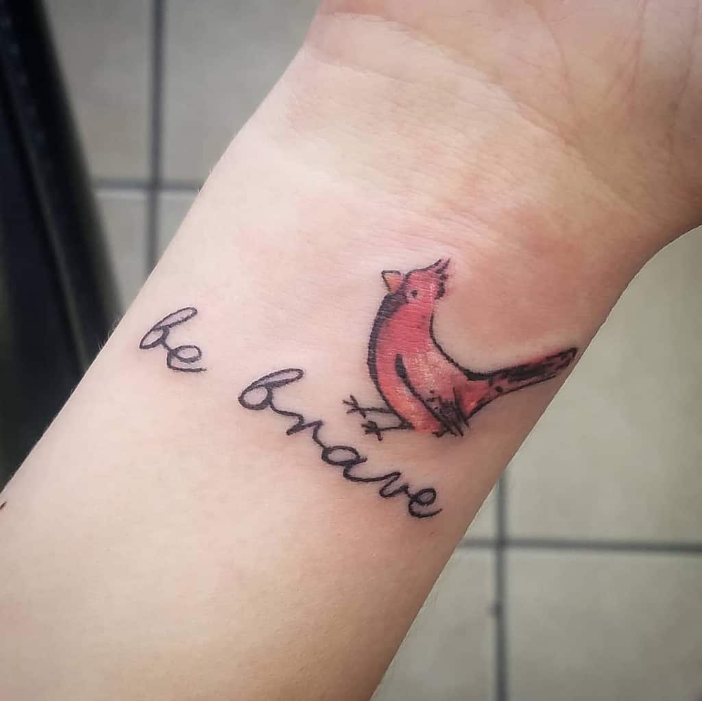 Wrist tattoo of a pink bird beside the words "be brave" in cursive