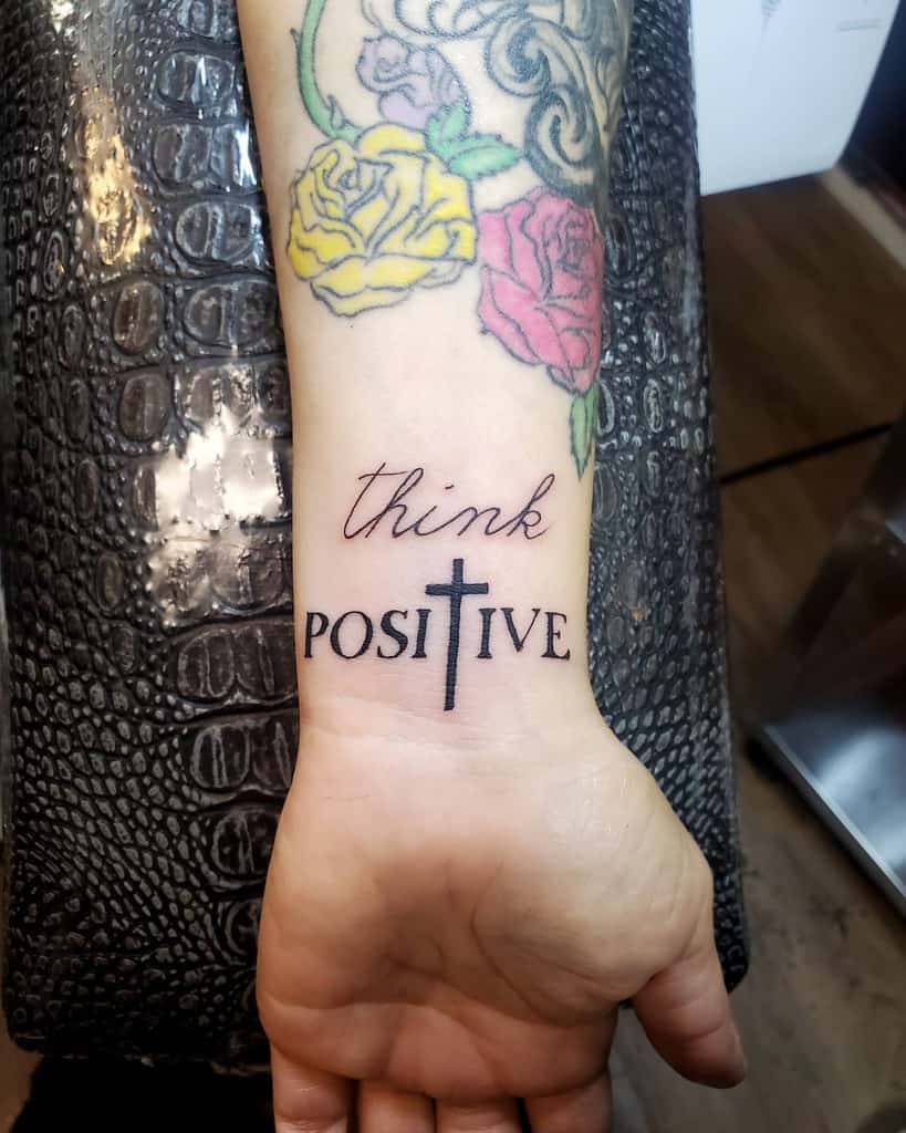 Wrist tattoo of "think positive" with a cross, surrounded by colorful floral designs