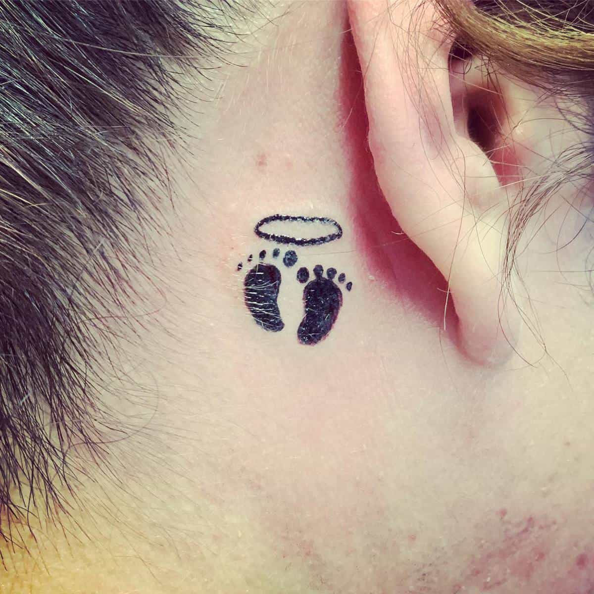 Tattoo of small footprints with a halo above them, located behind an ear