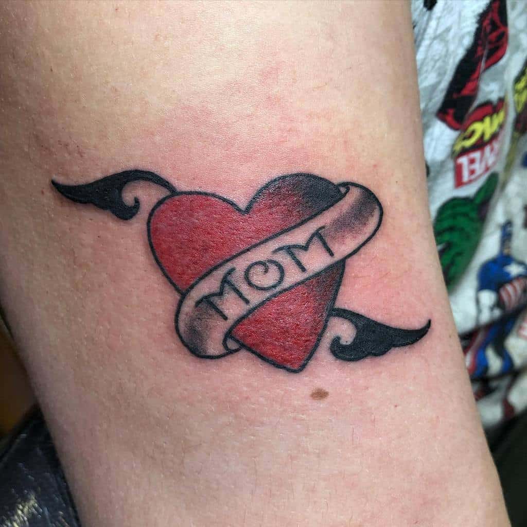Tattoo of a heart with wings and a ribbon that reads "MOM"