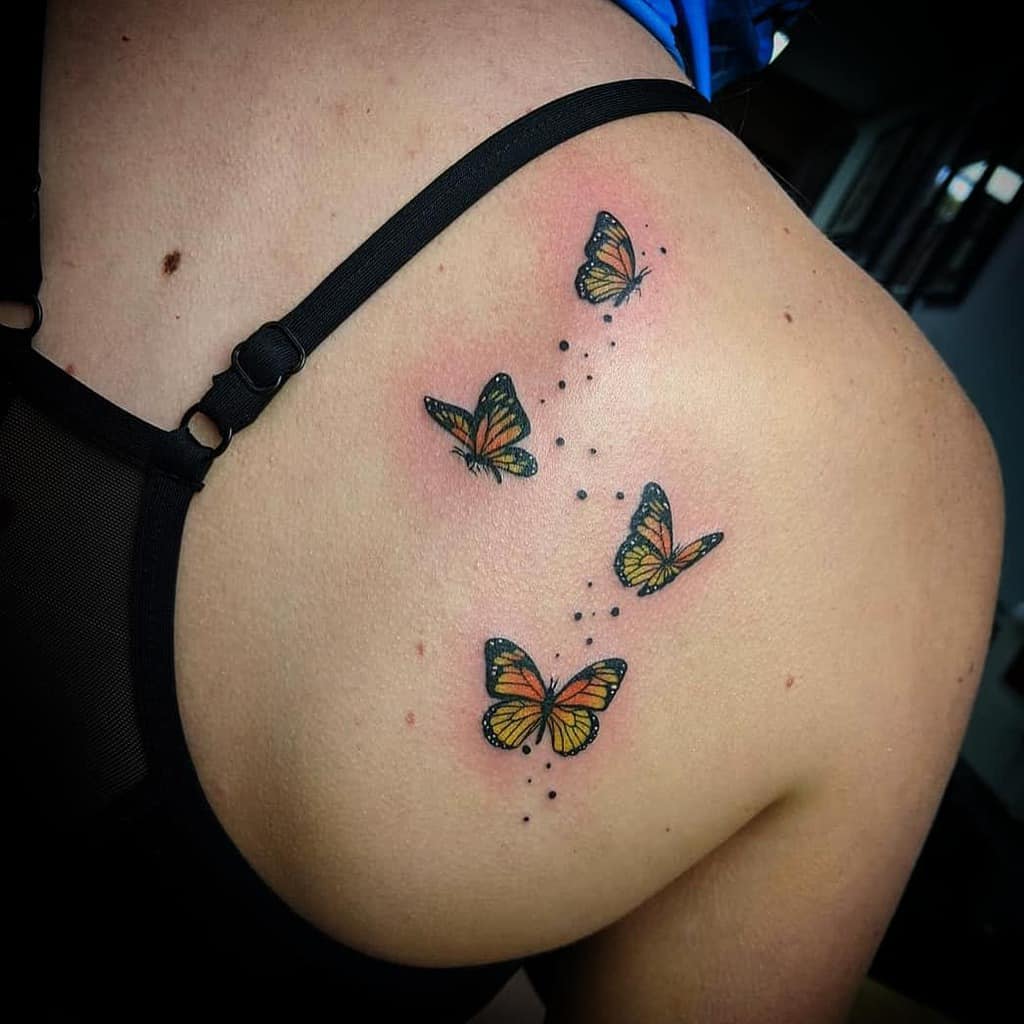 Tattoo of four butterflies on a person's upper back, resembling delicate monarchs in flight, with small black dots trailing them