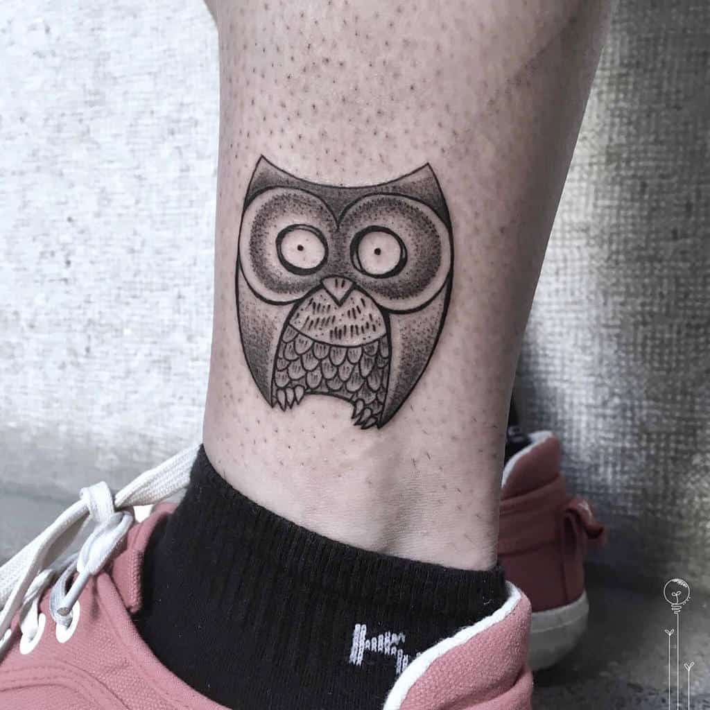 A quirky owl tattoo with oversized eyes and a geometric style, perched on the ankle in black and gray shading, offering a playful, minimalistic design