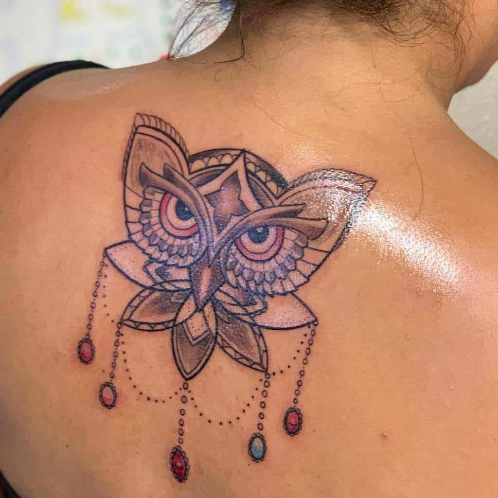 An owl tattoo with intricate geometric shapes and elegantly hanging jewels adorns a person's shoulder