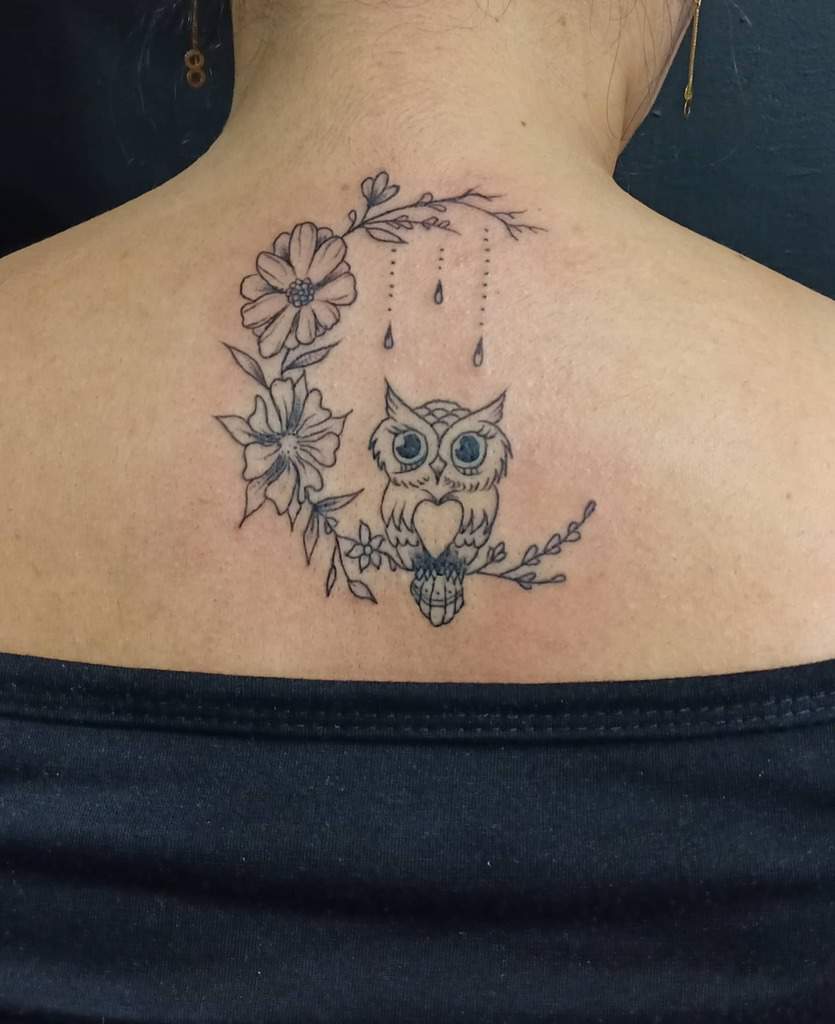 An intricate owl tattoo on the back is artfully intertwined with blooming flowers and delicate, dangling beads