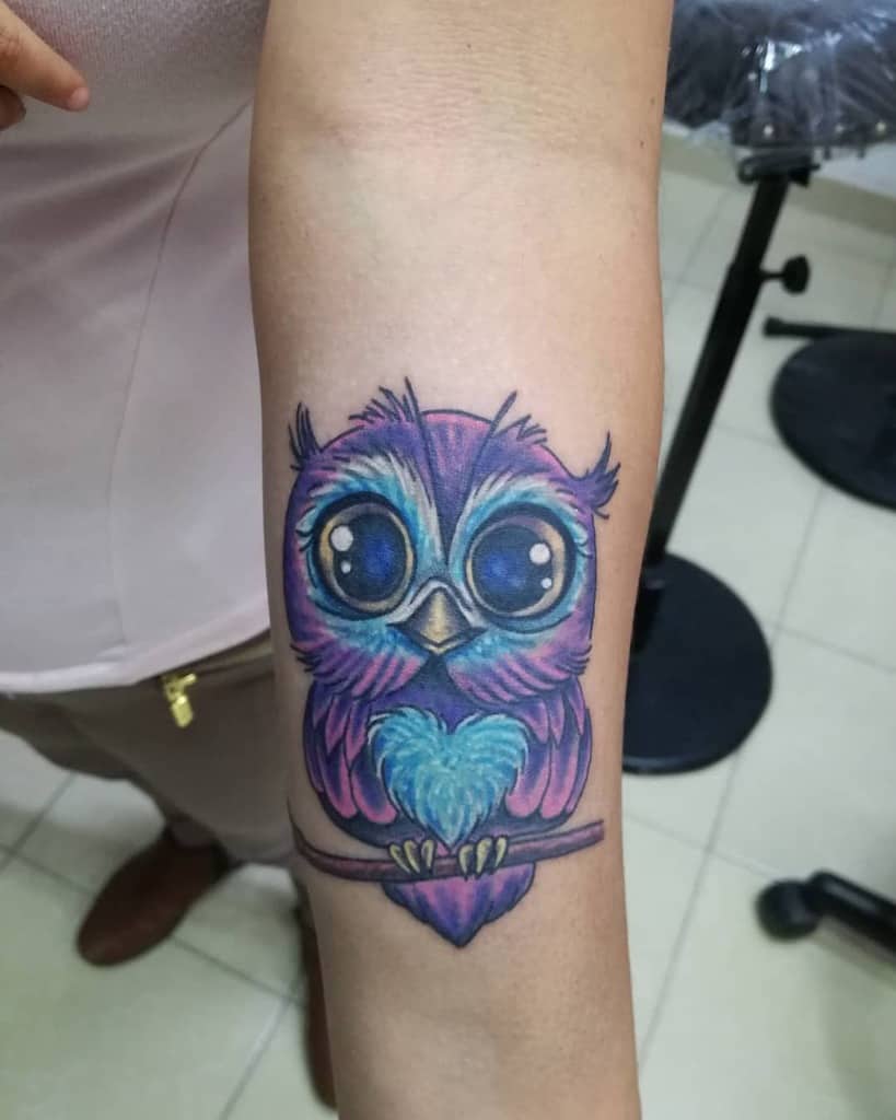 A vibrant owl tattoo adorns the forearm, showcasing its big eyes and an explosion of purple and blue hues