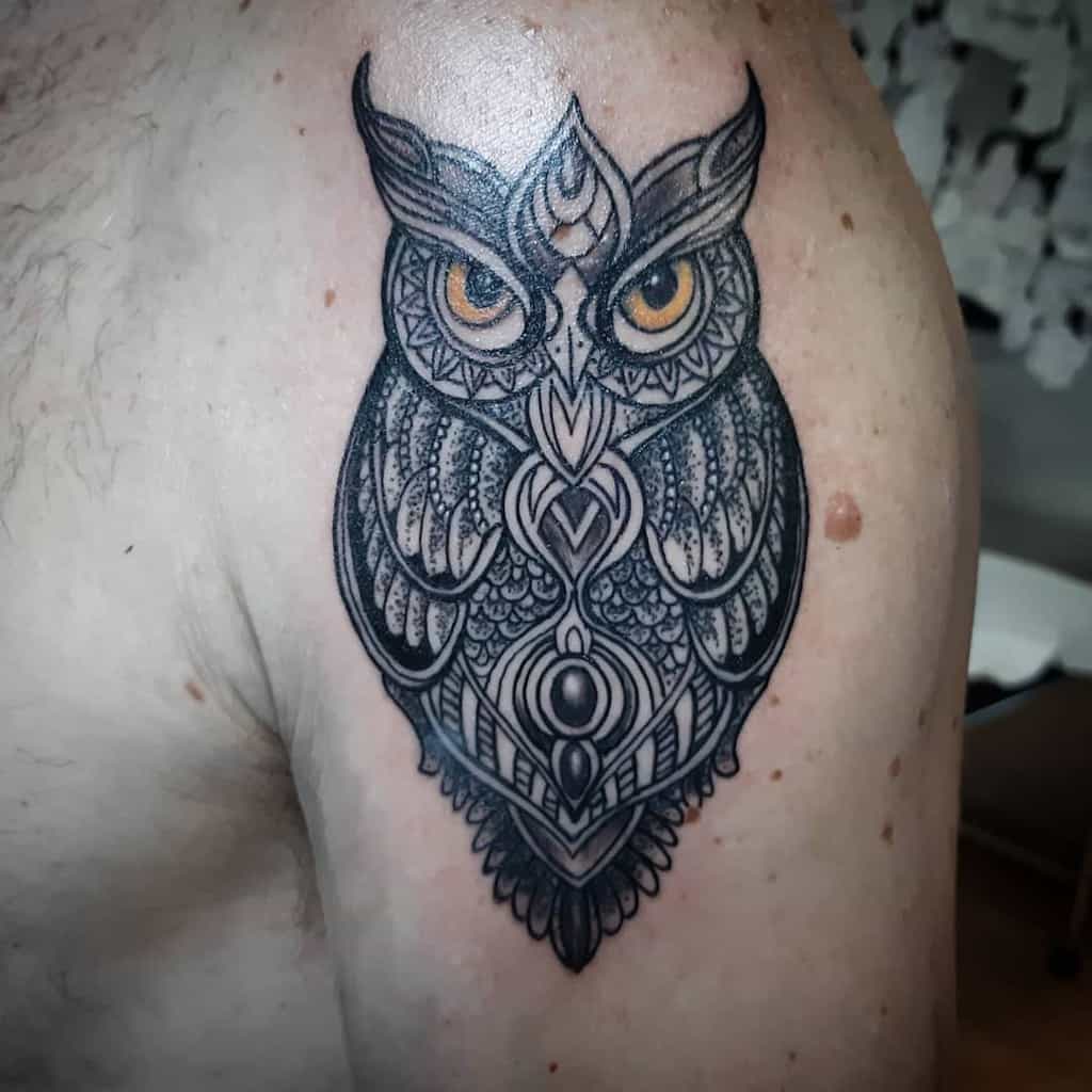 A geometric owl tattoo on the shoulder, featuring intricate linework and detailed patterns around the eyes, giving it a mystical appearance