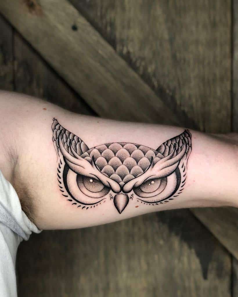 A detailed black ink tattoo of an owl with intricately designed feathers and large eyes, placed on the upper arm