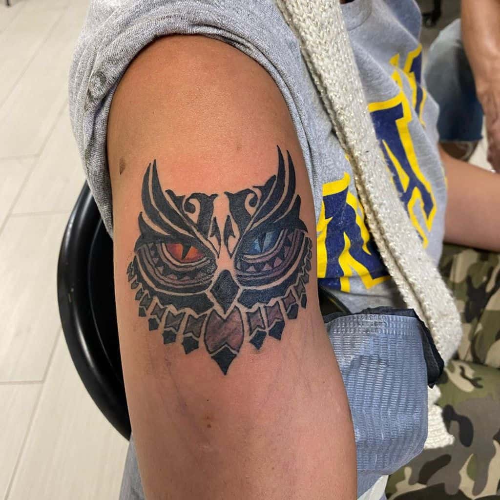 Owl tattoo with intricate patterns and blue and red eyes on a person's upper arm