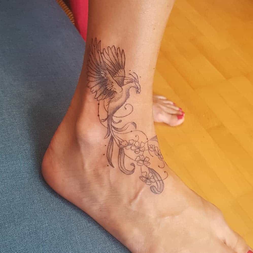 A detailed phoenix ankle tattoo in black ink, featuring flowing feathers and floral accents beneath