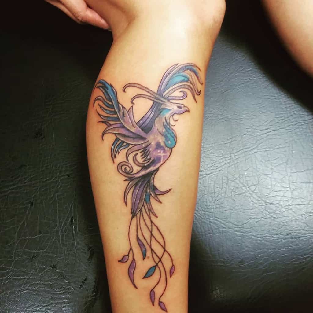 A vibrant phoenix tattoo on the leg, featuring a mix of purple, blue, and teal colors with flowing tail feathers