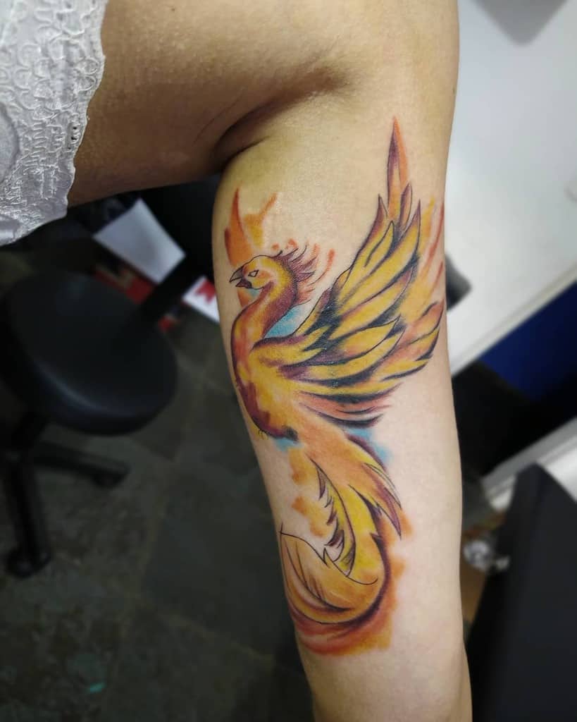 Colorful phoenix tattoo on inner arm featuring vibrant yellow, orange, and blue hues
