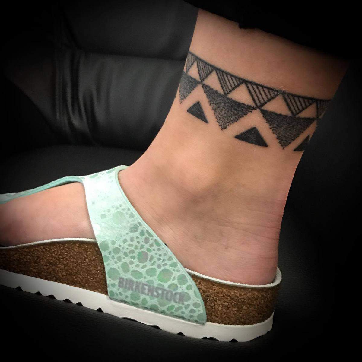 Close-up of an ankle with a black geometric tattoo, wearing a light green Birkenstock sandal