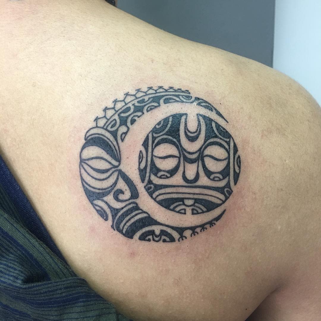 A shoulder tattoo featuring a circular design with intricate, tribal-style patterns and abstract faces