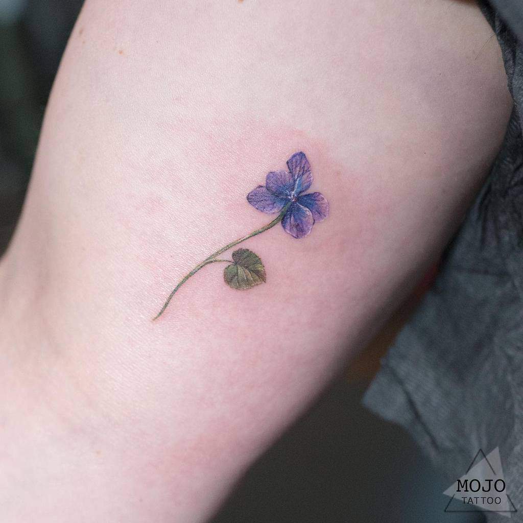 Small purple flower tattoo with a green leaf on the upper arm