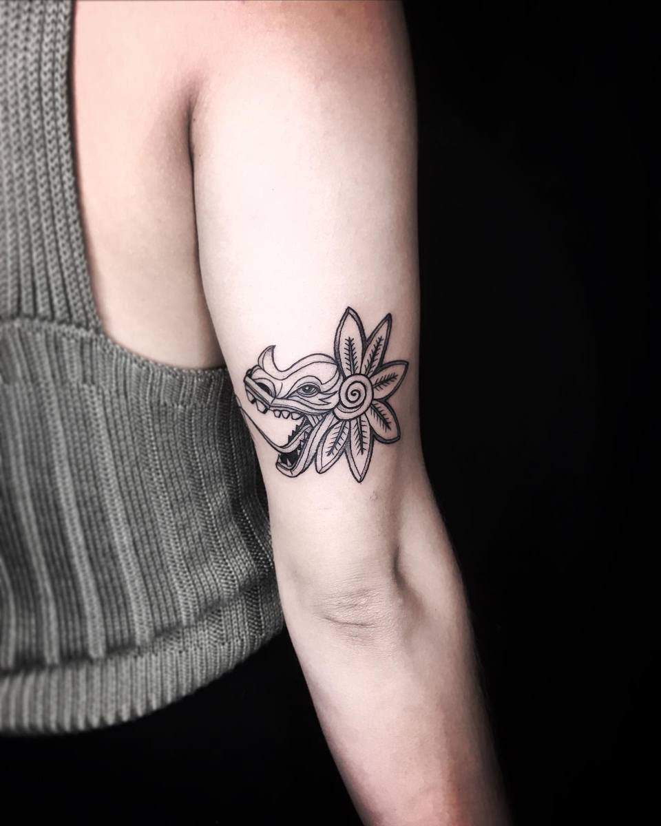 Minimalist upper arm tattoo of Quetzalcoatl's head with a stylized flower element