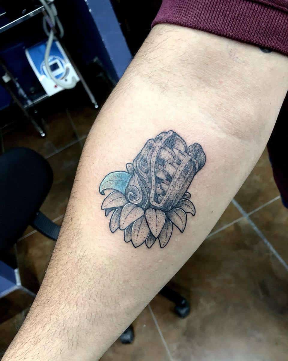 Small inner arm tattoo of Quetzalcoatl's head, featuring subtle blue shading and floral elements