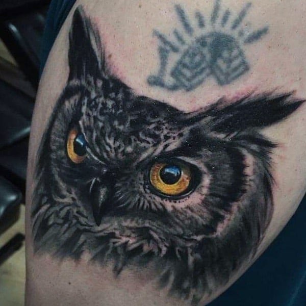A tattoo of a mesmerizing owl with vivid eyes sits gracefully below a crown-like symbol on a person's arm