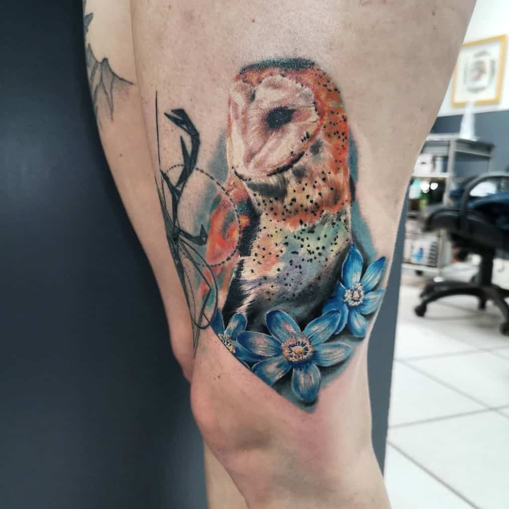 A vibrant owl tattoo perched amidst blue flowers adorns a person's leg, creating a stunning tapestry of color and nature