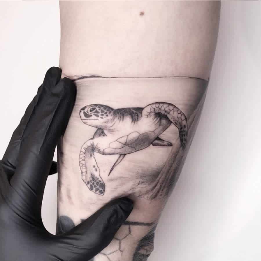 A realistic black and grey tattoo of a turtle swimming, located on the forearm, with a hand gently touching it