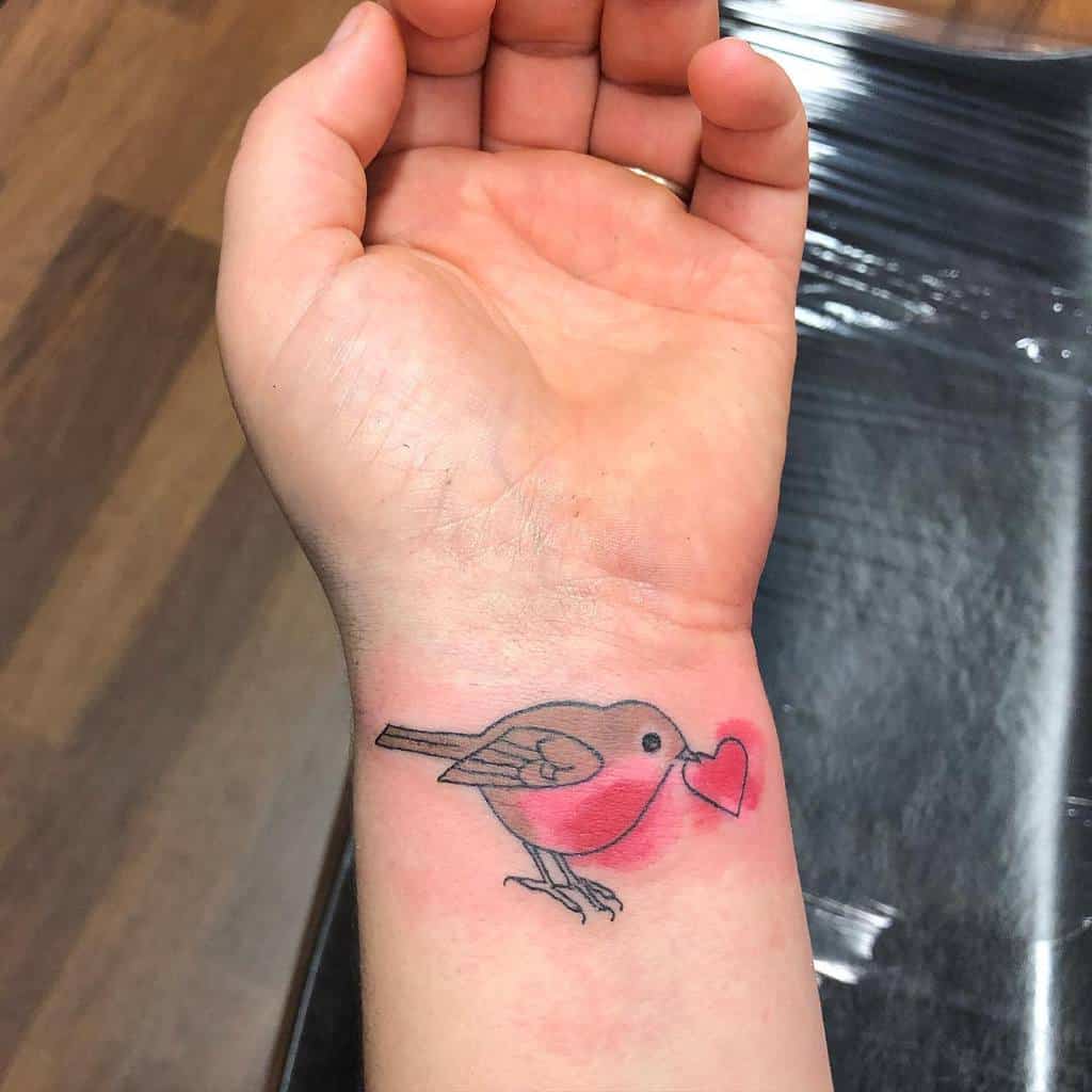 A small colorful tattoo of a robin with a red and pink chest and beak holding a red and pink heart