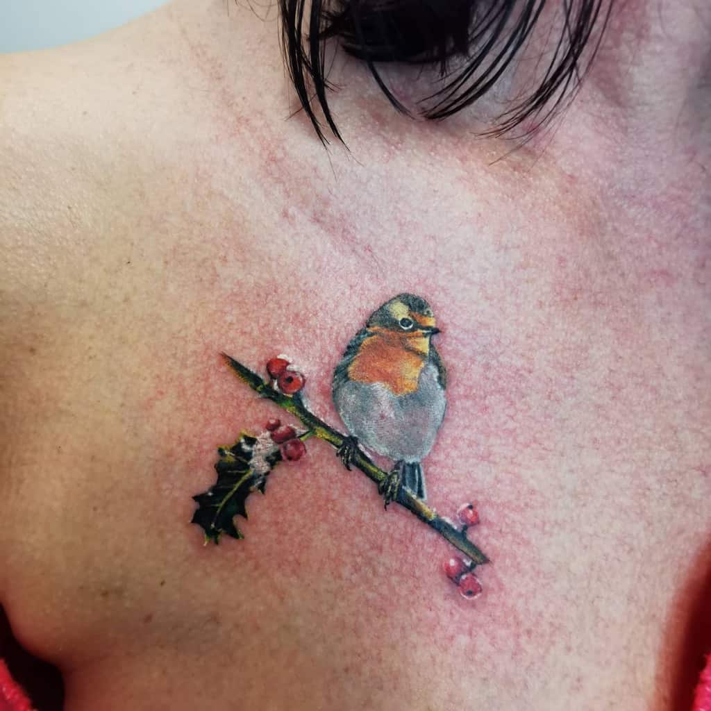 Realistic tattoo of a robin perched on a holly branch with red berries and one green leaf, featuring vibrant colors and fine shading on the robin’s chest and feathers