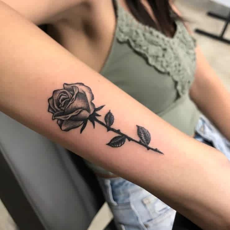 Tattoo of a detailed black rose with leaves on a person's forearm
