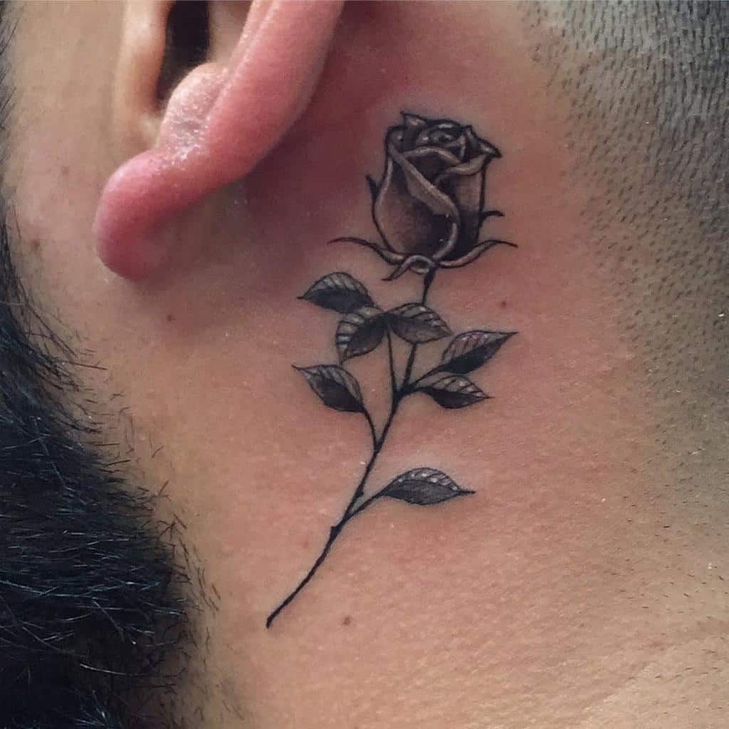 Black rose tattoo behind a man's ear with detailed leaves and stem