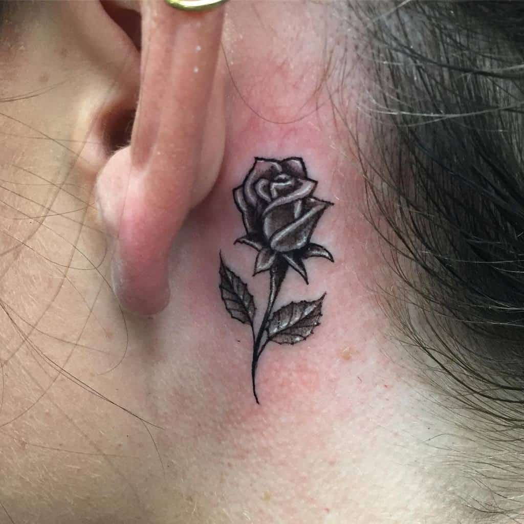 Black and grey rose tattoo behind the ear, featuring fine-line details and soft shading for a bold yet delicate look