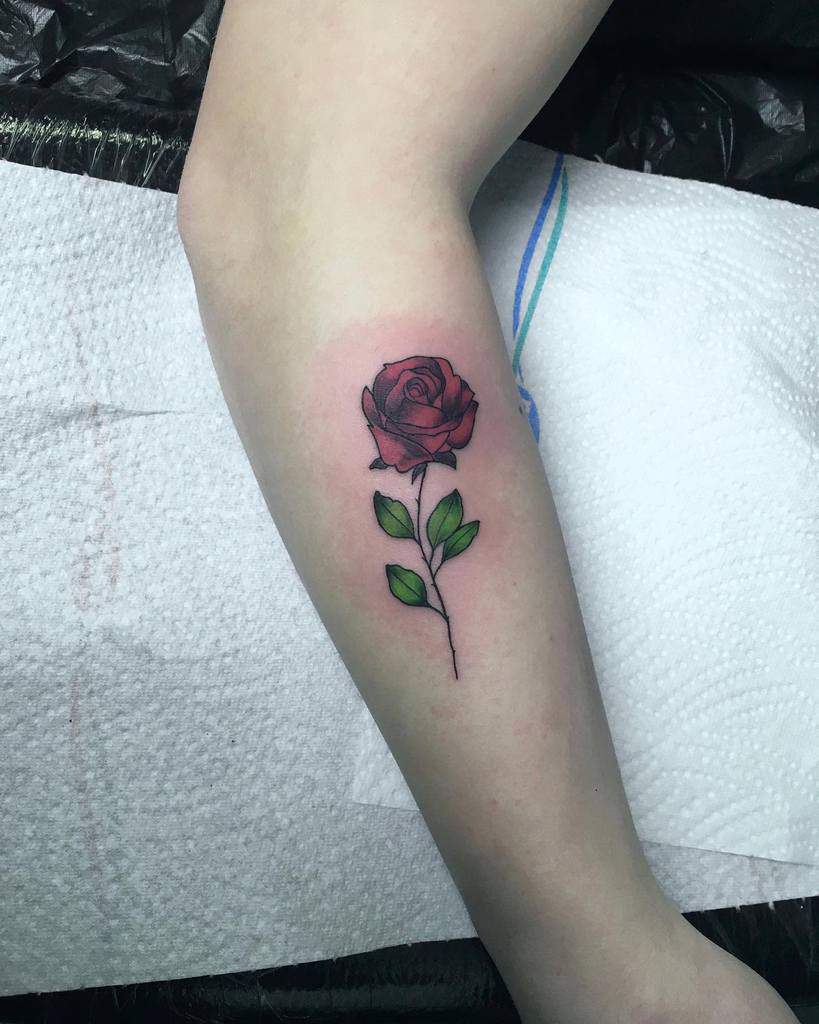 A red rose tattoo on a forearm with green leaves