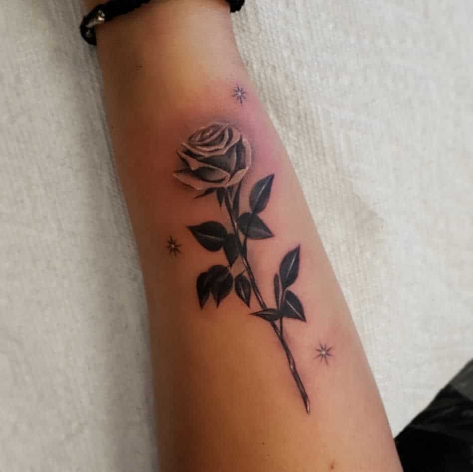 Forearm tattoo of a black and gray rose with leaves, surrounded by small stars on a forearm