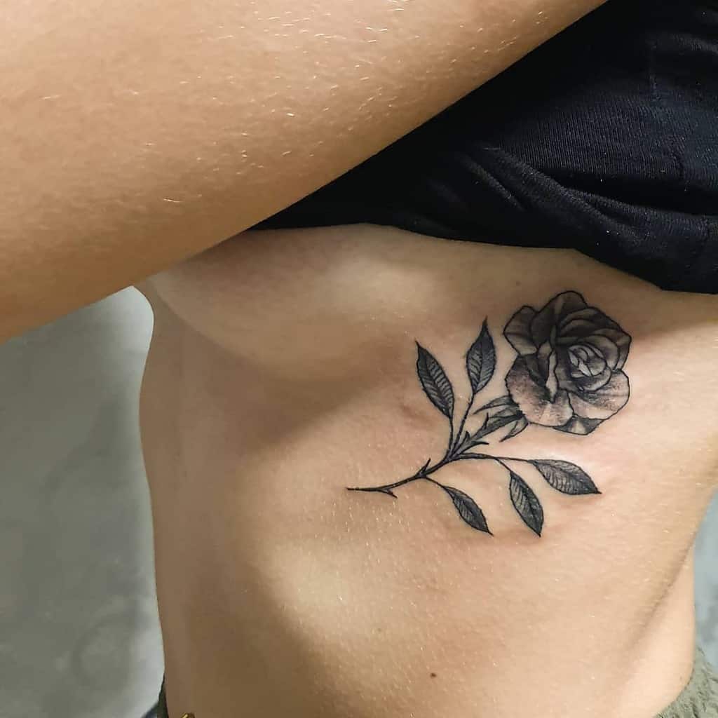A black and grey rose tattoo with leaves on woman's ribcage