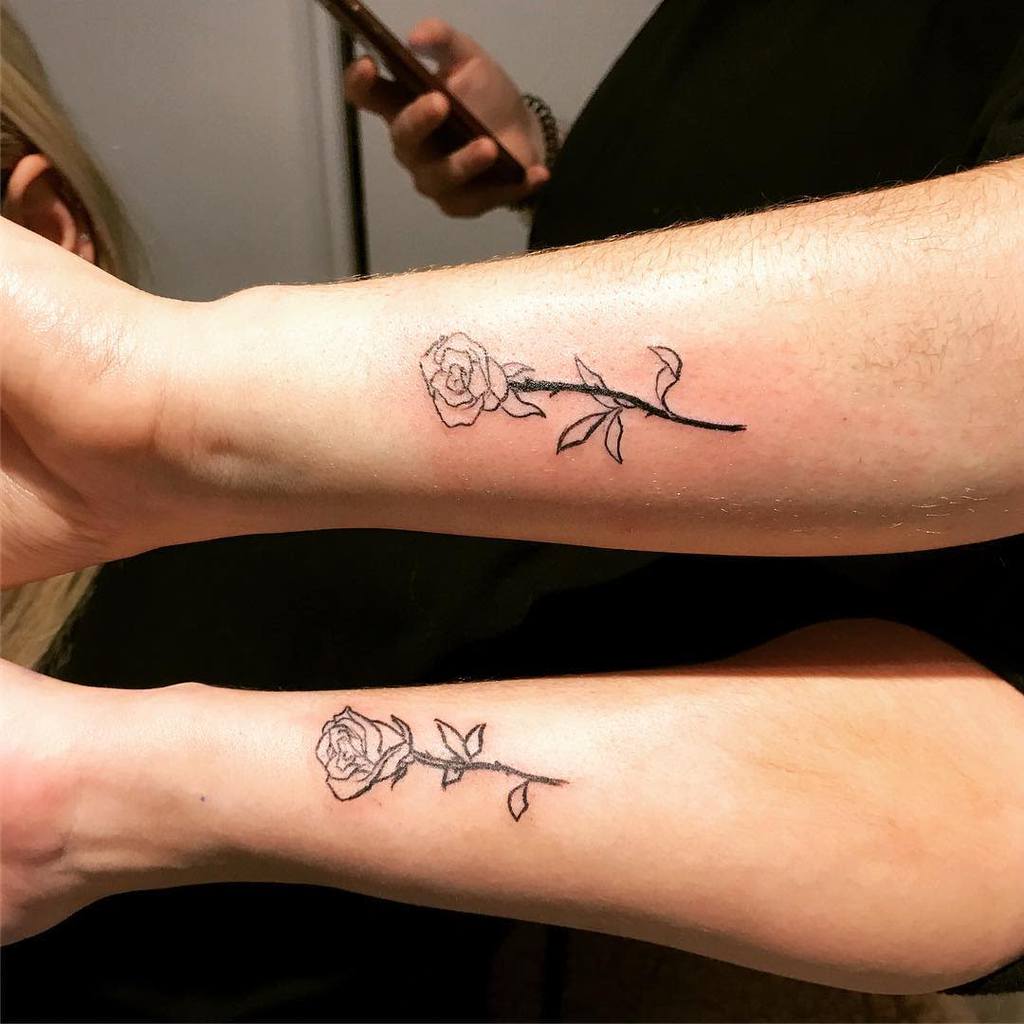 Two forearms with matching black line tattoos of roses with stems and leaves