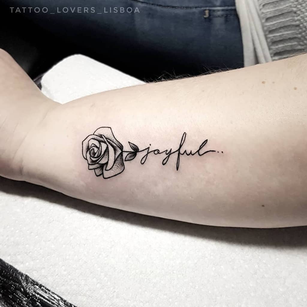 Tattoo of a rose with the word "joyful" on an arm