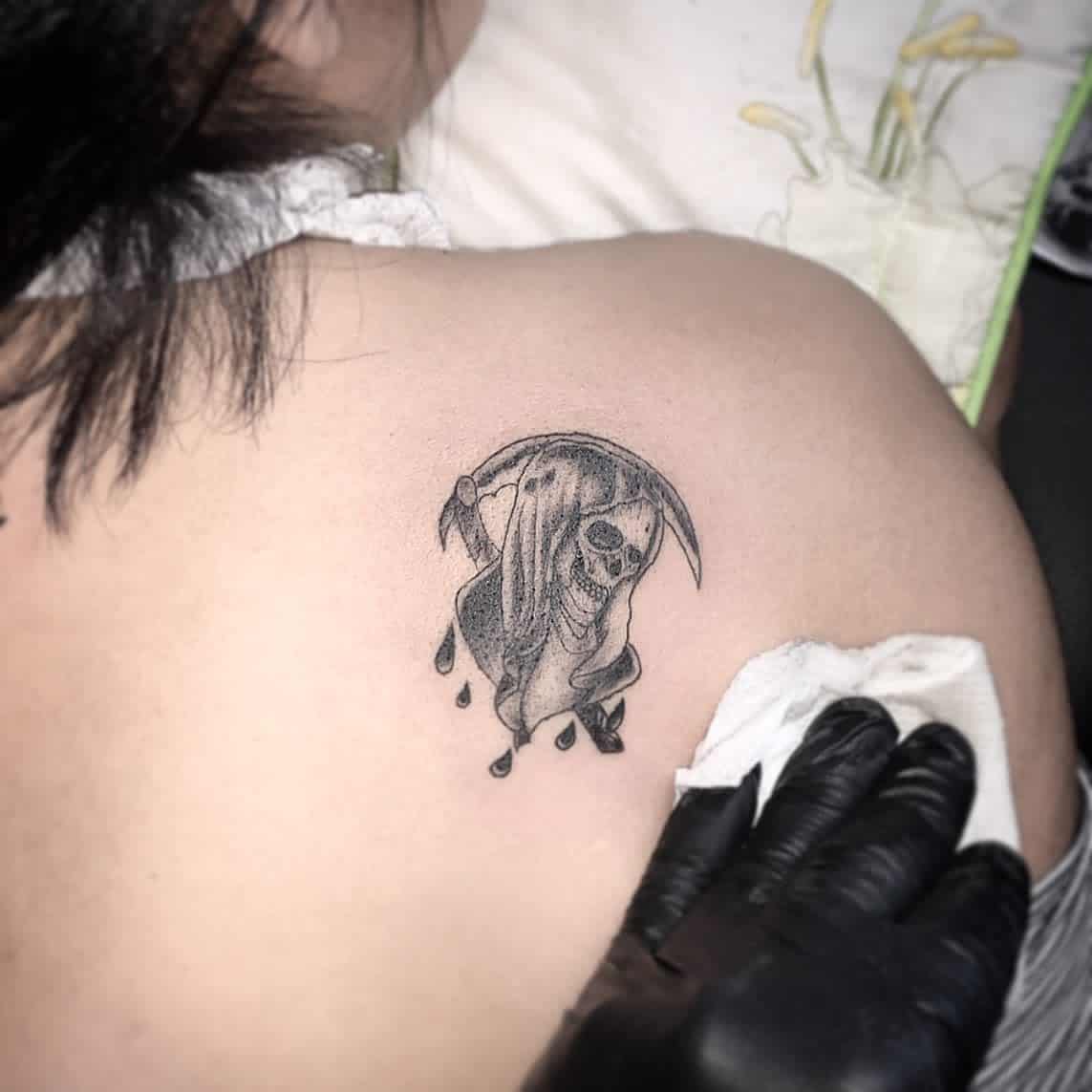 A small Santa Muerte tattoo located on the upper back, featuring a hooded skeleton holding a scythe, with droplets below, symbolizing protection and devotion in a subtle design