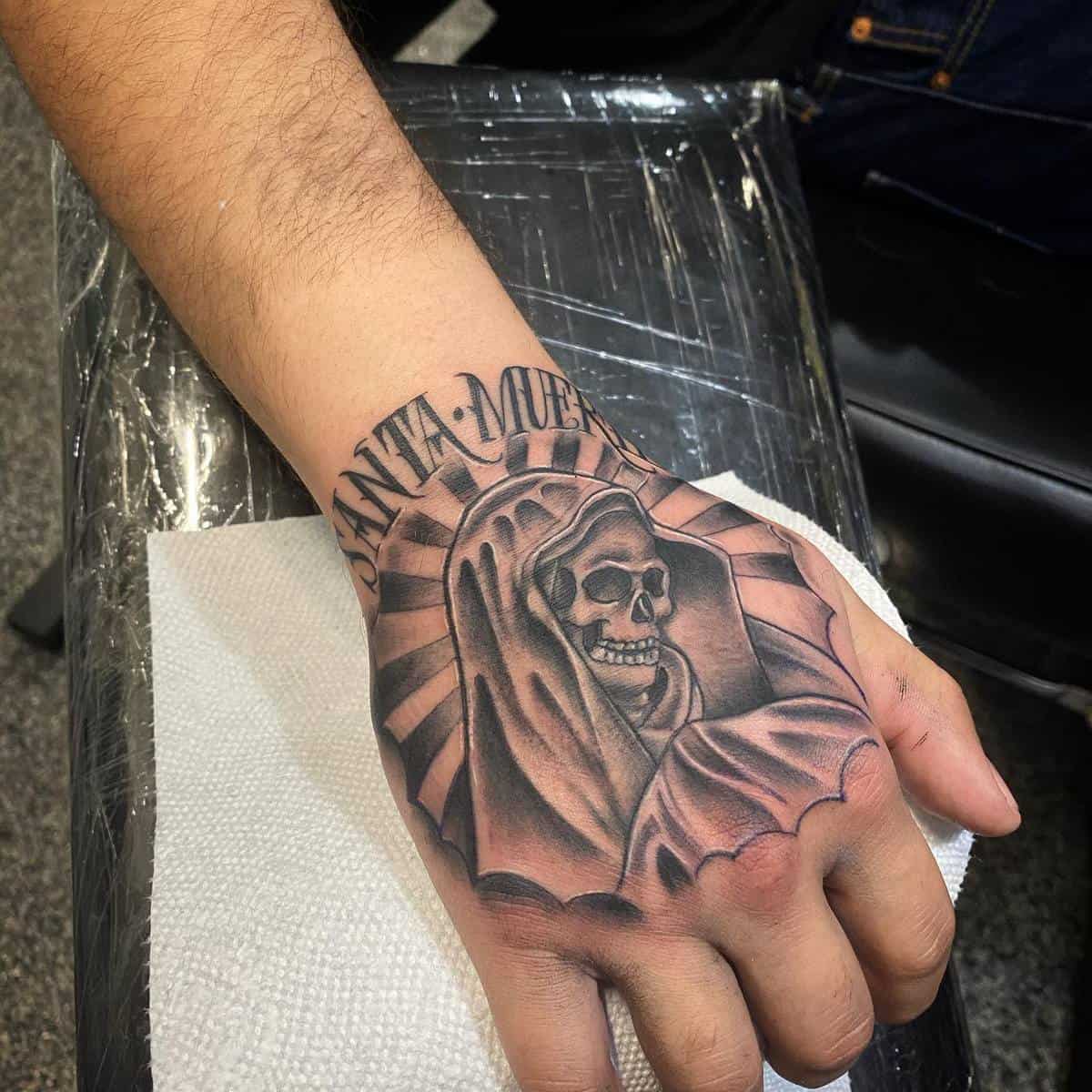 Santa Muerte hand tattoo with hooded skeleton, sun rays, and bold "Santa Muerte" lettering