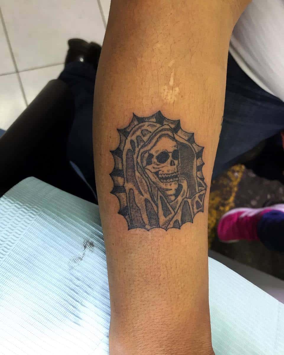 Small Santa Muerte tattoo featuring a hooded skeleton framed by a sunburst design on the forearm