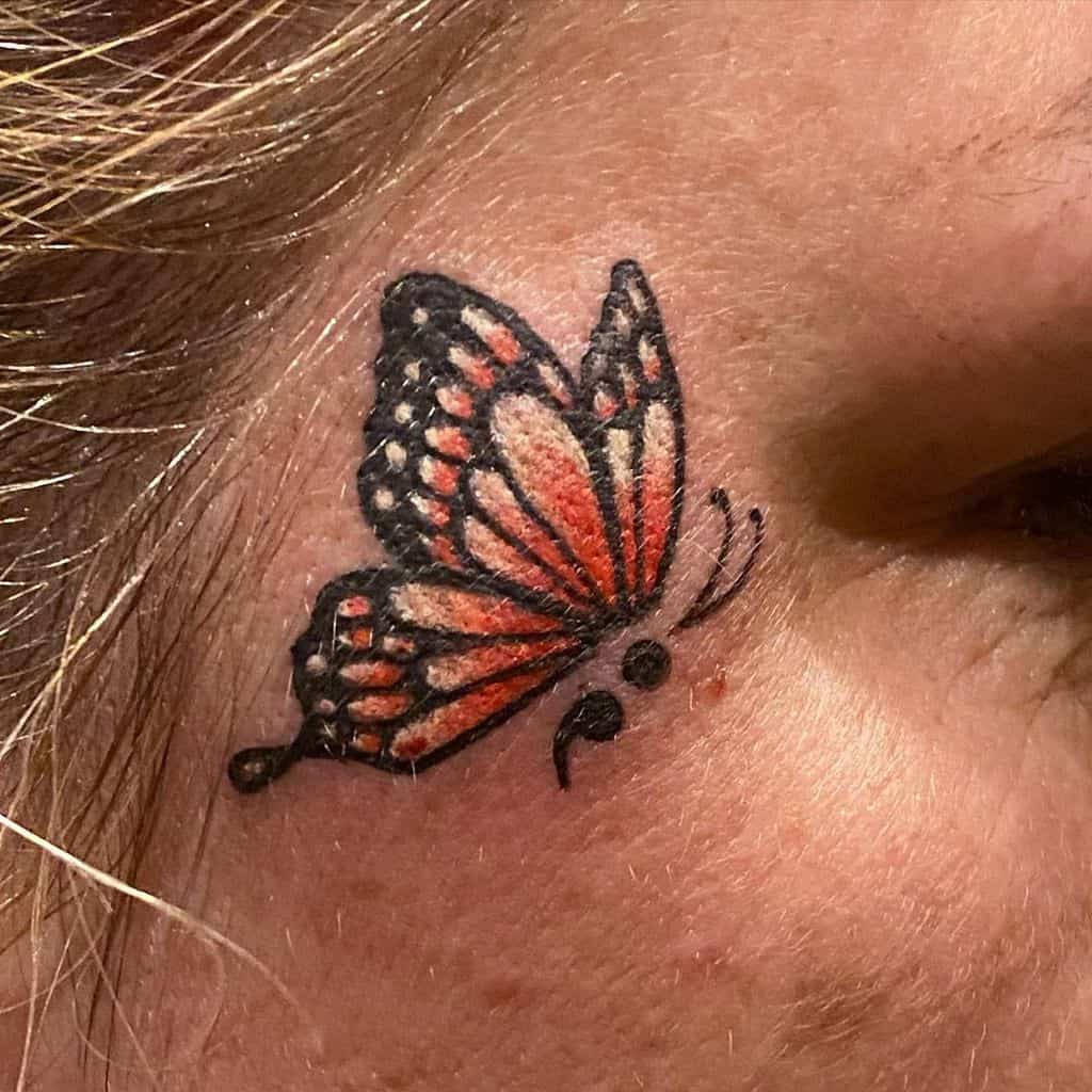 Semicolon butterfly tattoo, delicately placed near the eye, features vibrant orange and black wings