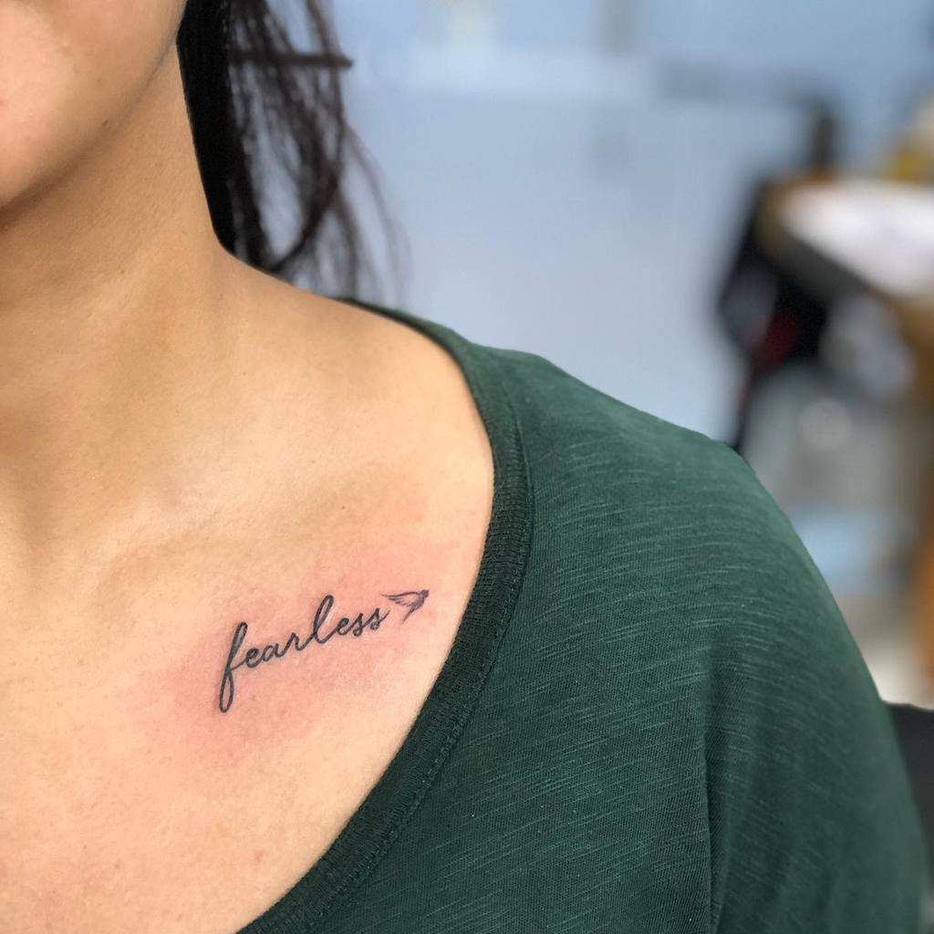 A tattoo of the word "fearless" with an arrow is on a woman's upper chest