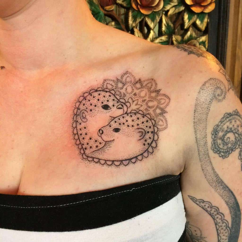 Delicate tattoo of two otters forming a heart shape, with intricate dot shading and lace-like mandala details on the chest