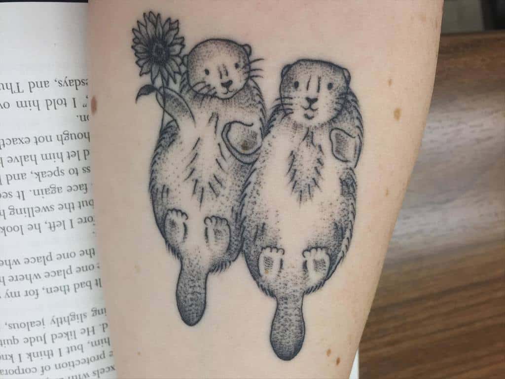 Tattoo of two playful otters holding hands, one clutching a sunflower, beautifully inked on an arm