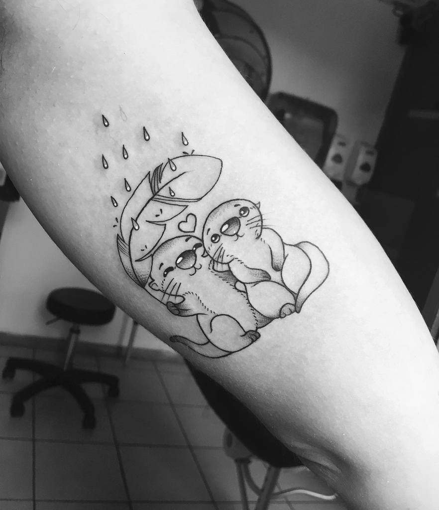 Black and white tattoo of intertwined otters with a heart above, sheltered by a leaf from raindrops
