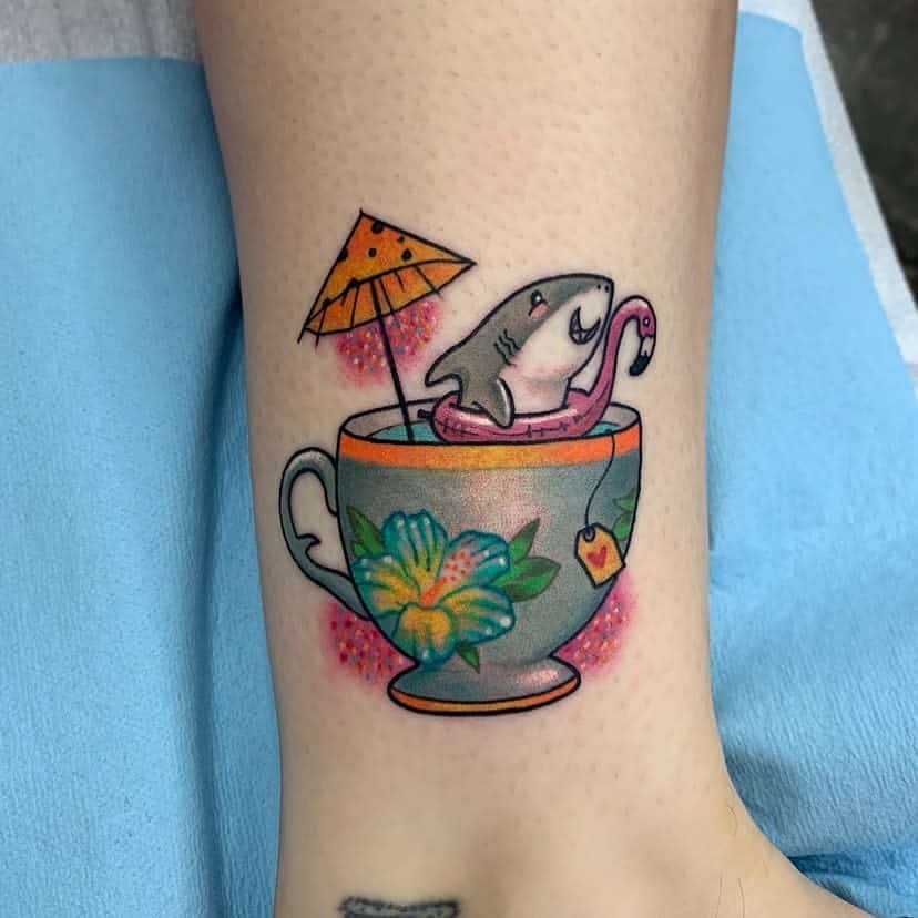Colorful tattoo of a shark in a floral design teacup with a tiny umbrella, flamingo floatie and a tea bag tag with a heart