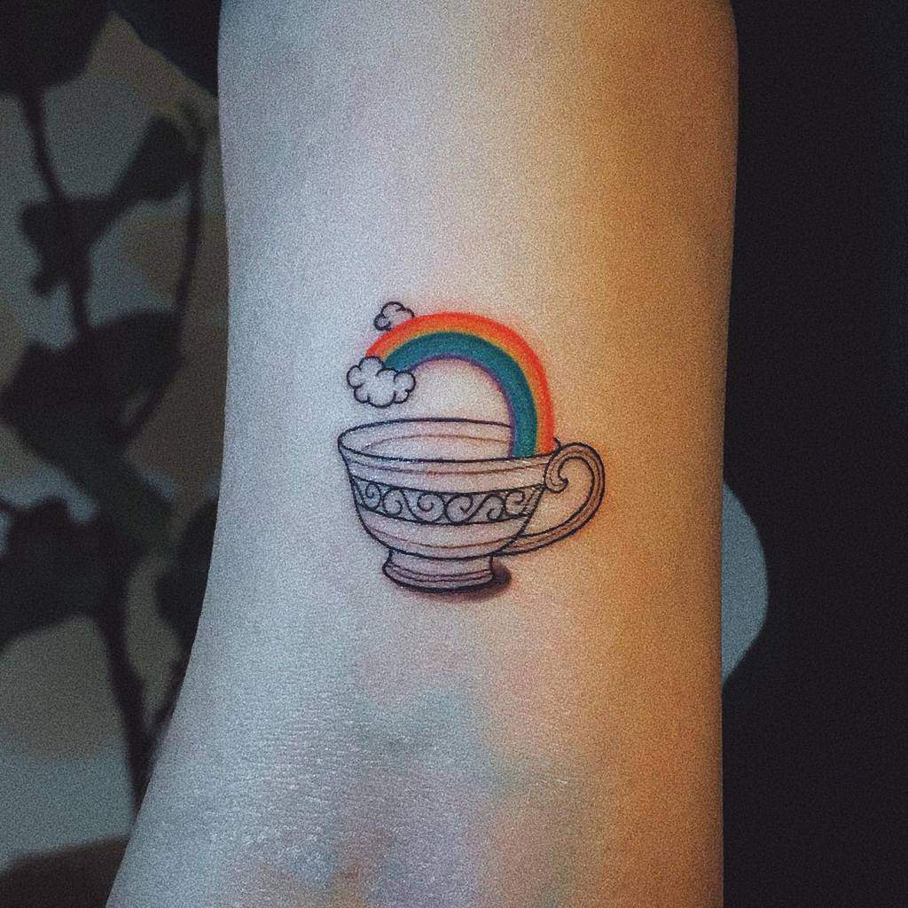 Tattoo of a teacup with swirling designs, filled with a rainbow and clouds