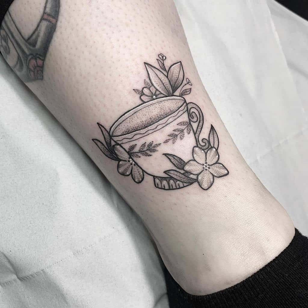 Black and gray tattoo of a teacup with floral designs and leaves 