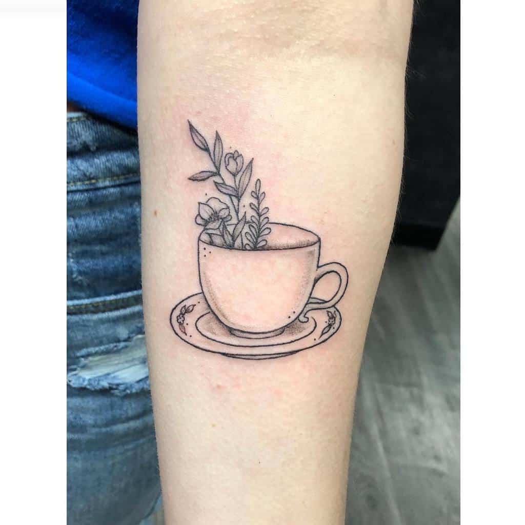 Minimalist black and grey tattoo of a teacup on a saucer, with delicate flowers and leaves growing from inside