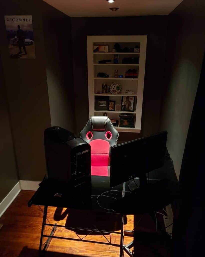 A dark gaming setup with a red and black chair, computer, and dual monitors on a desk in a small room