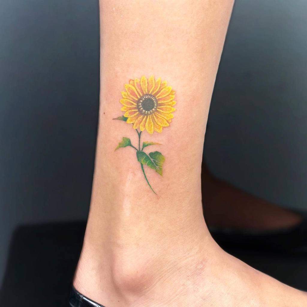 A small sunflower tattoo on an ankle, featuring a yellow bloom and green leaves