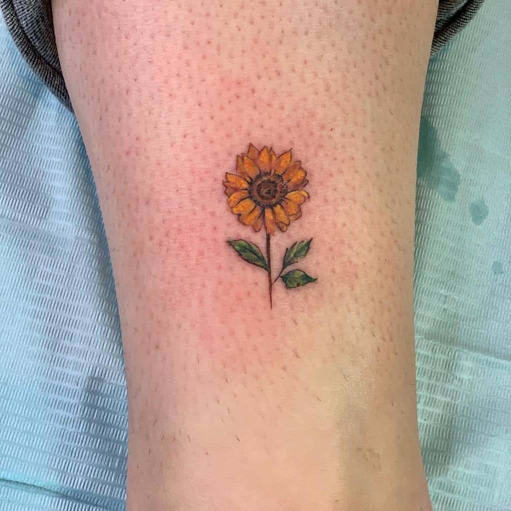 Small sunflower tattoo on an ankle with green leaves 