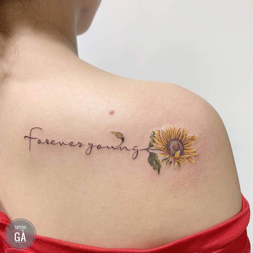 Minimalist tattoo on the shoulder with 'forever young' script and a delicate yellow sunflower design